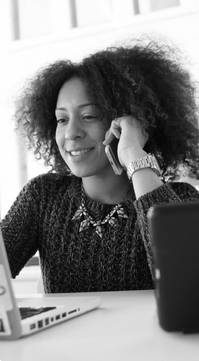 Banner em preto e branco com uma mulher sorridente, de cabelos crespos, com um casaco de lã escura e um relógio no pulso. Sentada a mesa, usa um notebook e um celular.